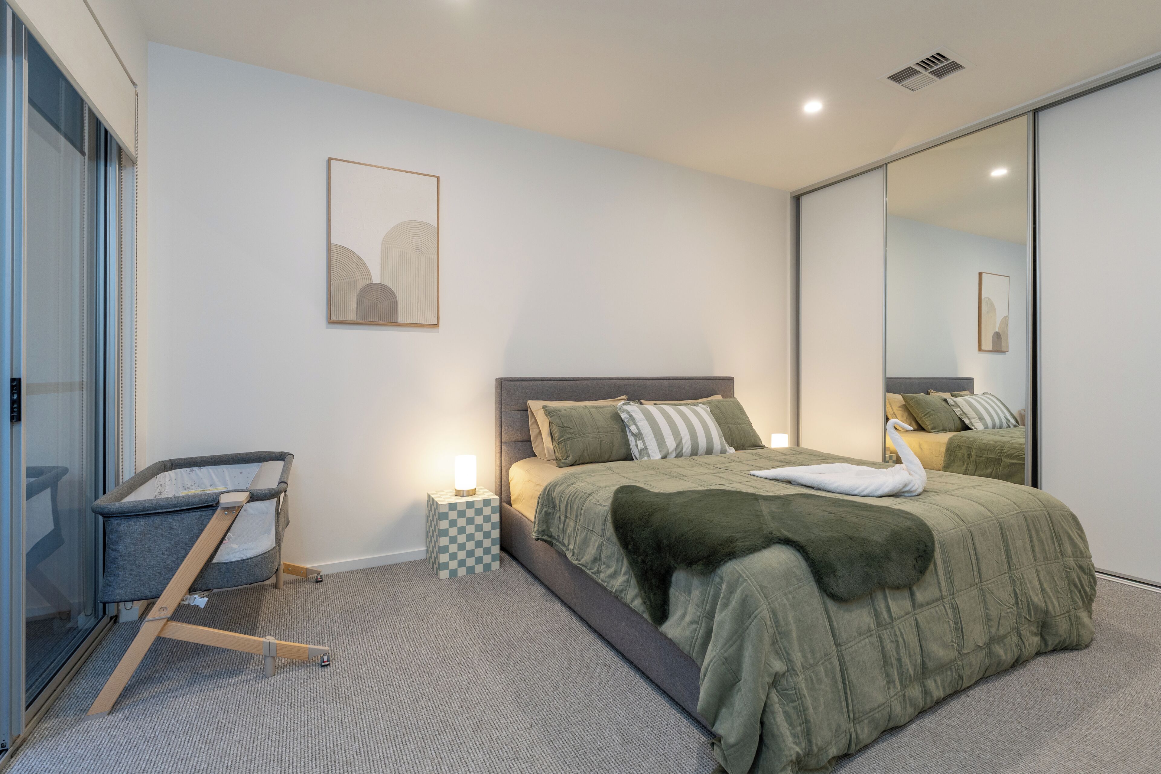 A modern bedroom with a bed, a baby crib, a mirror, and a framed picture on the wall.