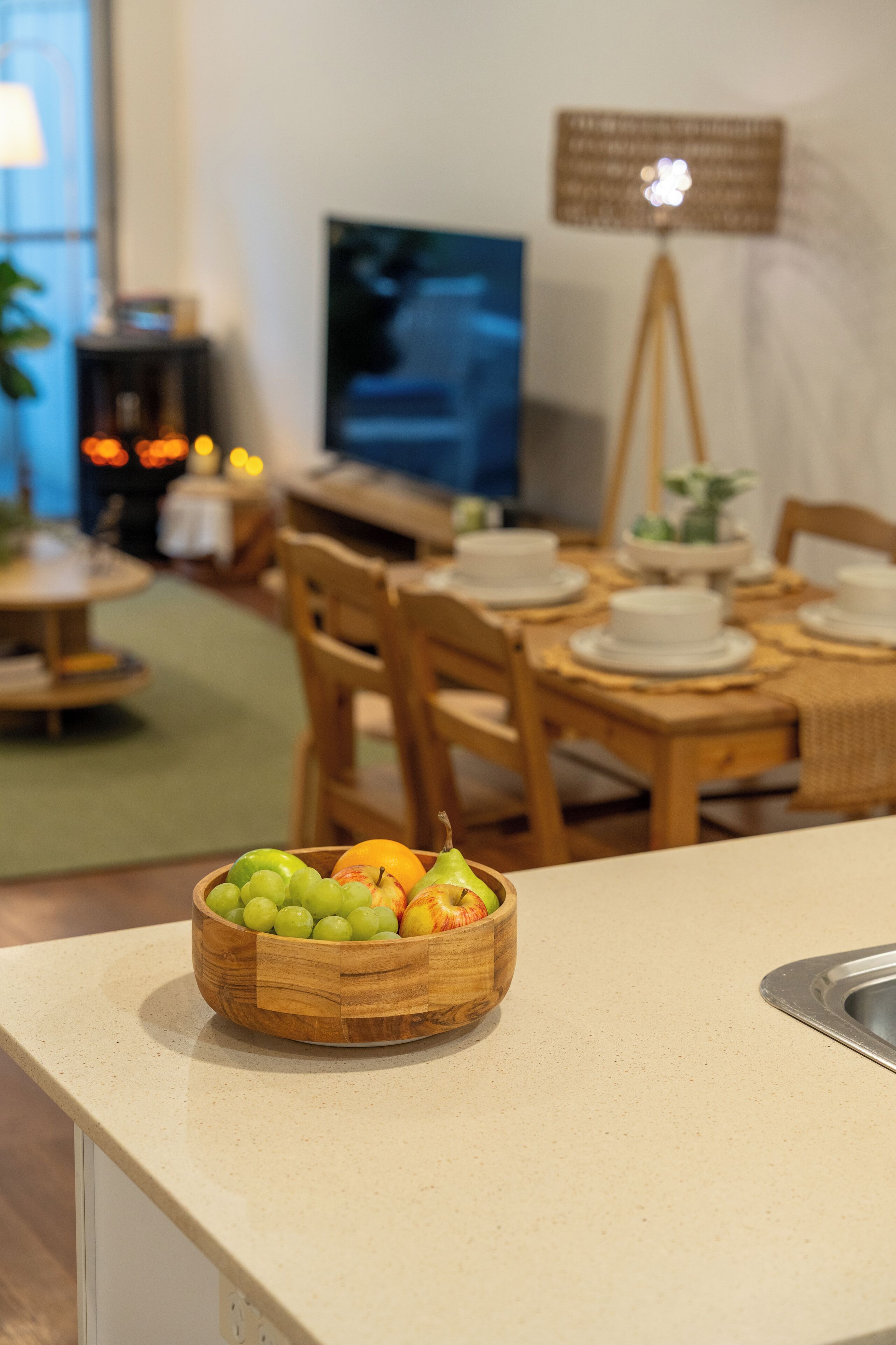 A kitchen counter with a fruit bowl, a dining table set for four, and a television in the background.