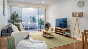 Ein modernes Wohnzimmer mit Sofa, Couchtisch und Fernseher. Es gibt einen Balkon mit Gartenmöbeln und Pflanzen.