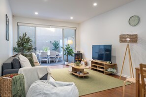 Ein modernes Wohnzimmer mit Sofa, Couchtisch und Fernseher. Es gibt einen Balkon mit Gartenmöbeln und Pflanzen.