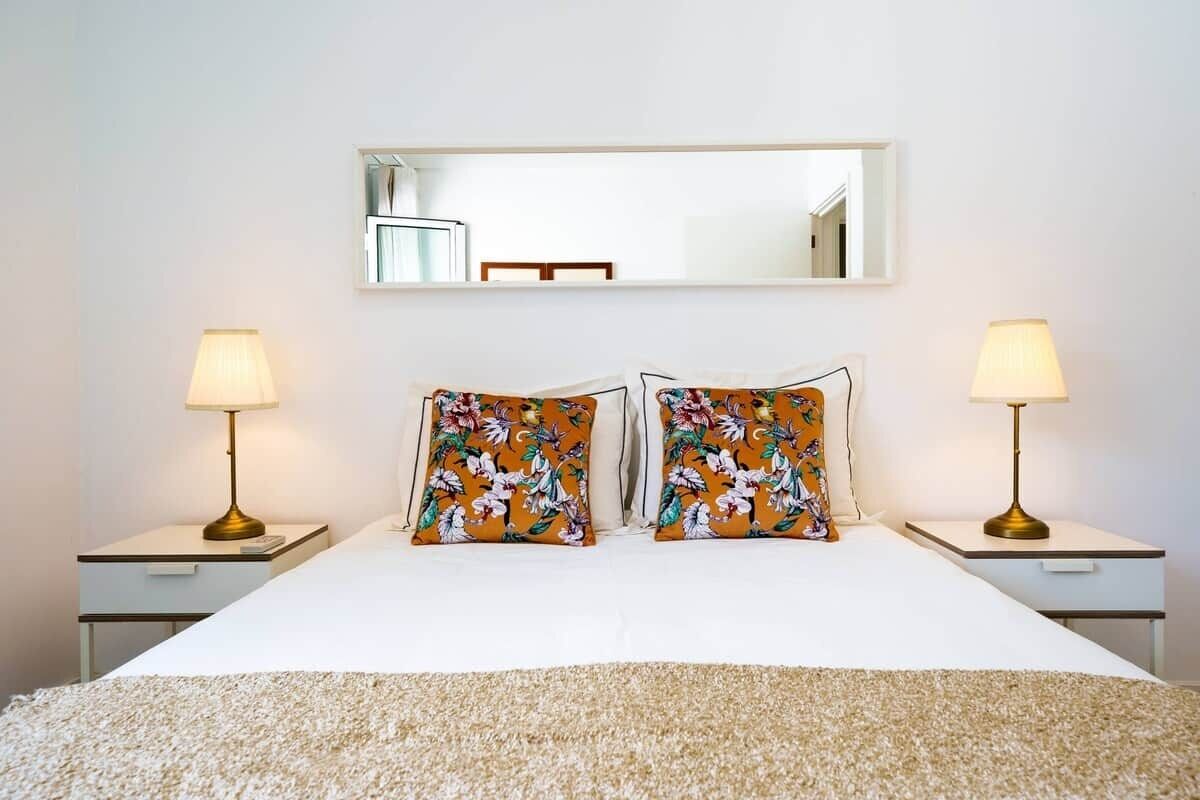 Un lit bien fait, agrémenté de coussins à motifs floraux, deux lampes de chevet et un miroir au mur.