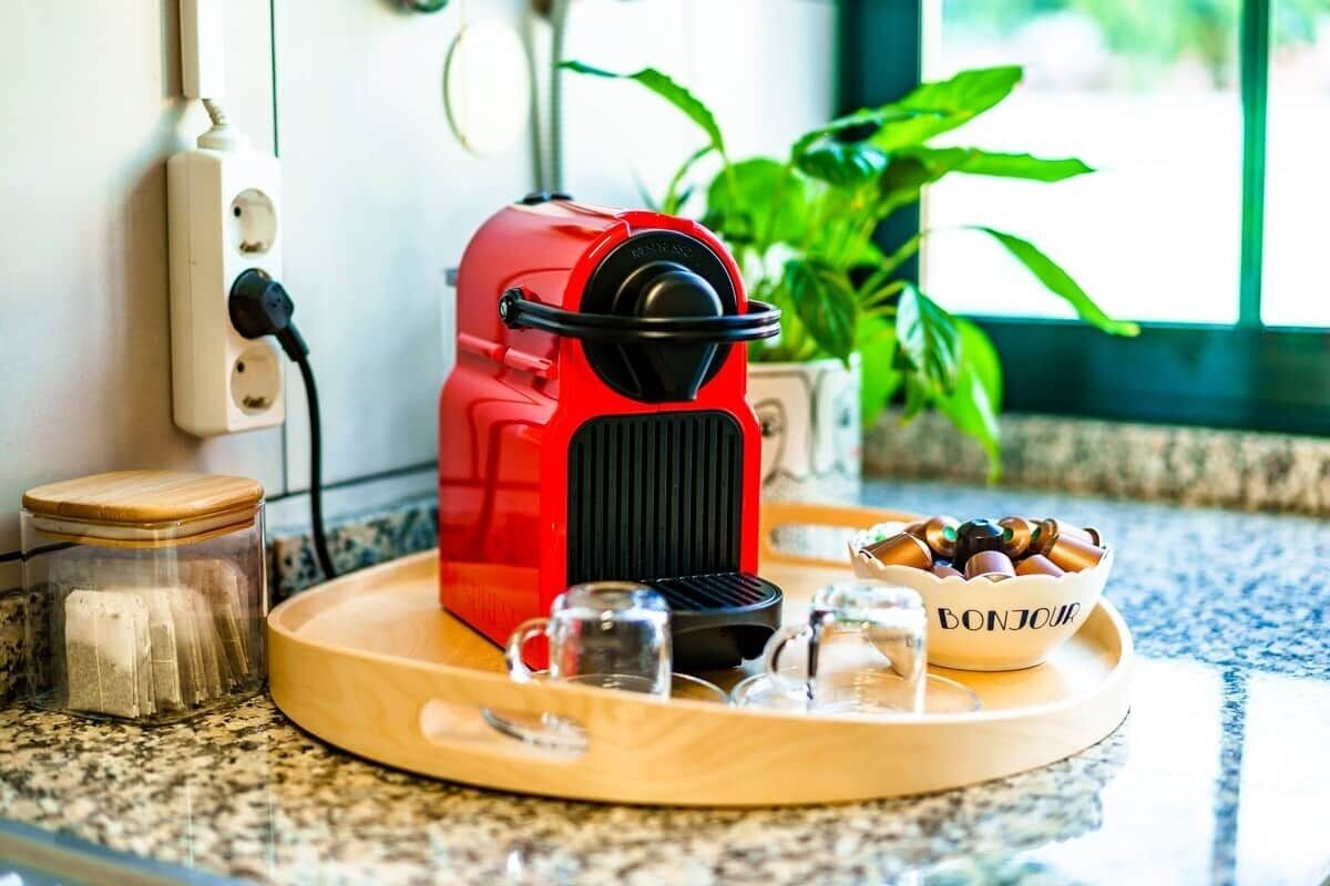 A red coffee maker on a wooden tray with a glass jar and a bowl of coffee beans.
