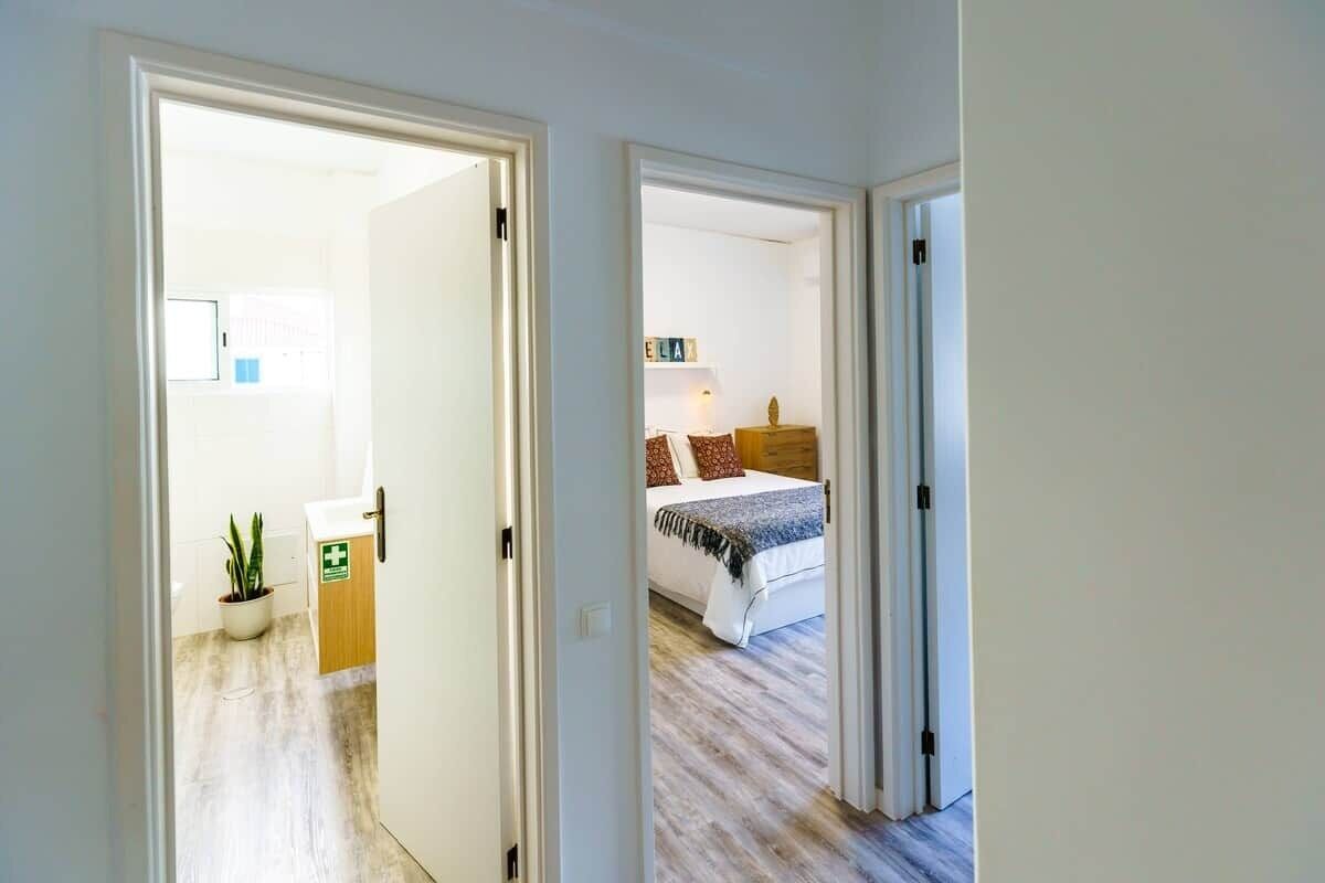 Un couloir mène à une chambre à coucher qui contient un lit, une table de chevet, une lampe et une petite plante.