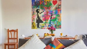 Ein Wohnzimmer mit grauem Sofa, weißen und bunten Kissen, einem Holzstuhl, einem Tisch mit Obstschale und Blumenvase sowie einem farbenfrohen abstrakten Gemälde an der Wand.