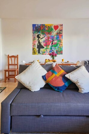 A living room with a grey sofa, white and multicolored pillows, a wooden chair, a table with a fruit bowl and a vase of flowers, and a colorful abstract painting on the wall. - Modern apartment with pool and garage (Lisbon)