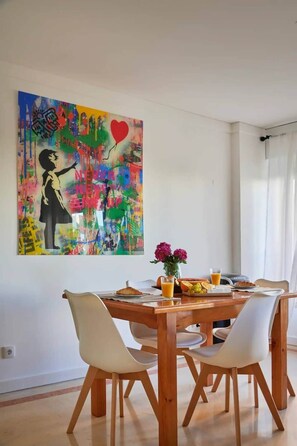 A dining area with a wooden table, white chairs, a colorful abstract painting, and a vase with flowers. - Modern apartment with pool and garage (Lisbon)