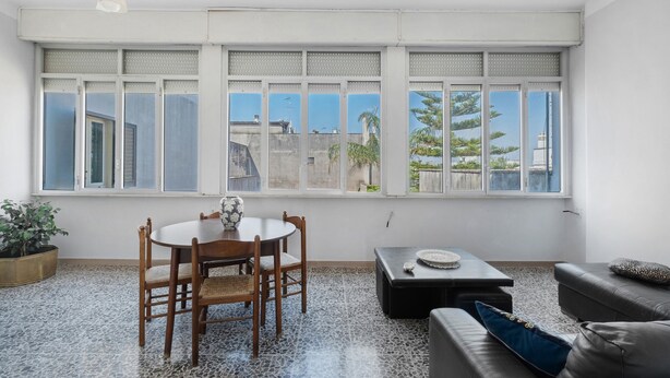 A room with a dining table, chairs, a sofa, a coffee table, and large windows with a view of buildings and trees.