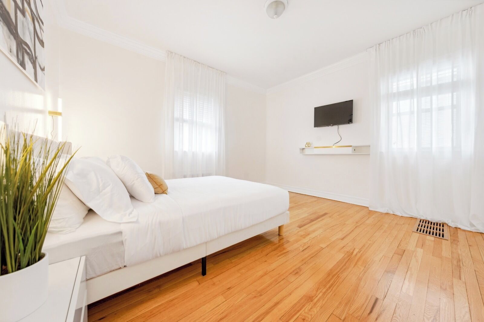 A bedroom with a bed, white curtains, a mounted TV, and a wooden floor.