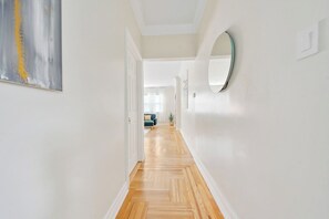 Un pasillo con suelo de madera, paredes blancas, un espejo redondo y una puerta que da a una habitación con sofá azul.