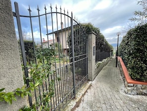 Een verhard pad dat leidt naar een poort met een hek van metaal, omgeven door groen en een stenen muur.
