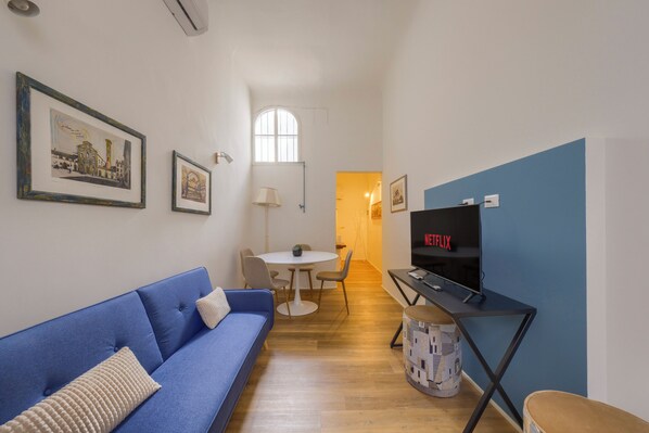 A compact living space with a blue sofa, a dining table with chairs, a television, and framed pictures on the walls. - Cellini's Historic Apartment • Historic Center • 5 min to the Duomo •10 min from the station (Firenze)