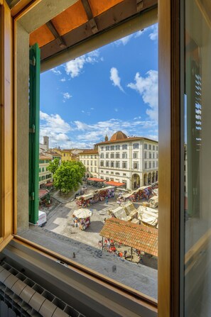 Blick aus einem Fenster eines Gebäudes mit einem Ziegeldach, einem großen Baum und einem Marktplatz mit Zelten und Menschen.