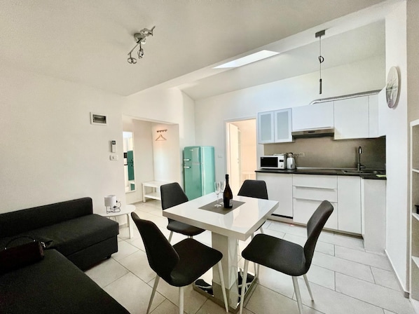 A compact living space with a dining table, black chairs, a black sofa, a kitchenette, and a teal-colored refrigerator.