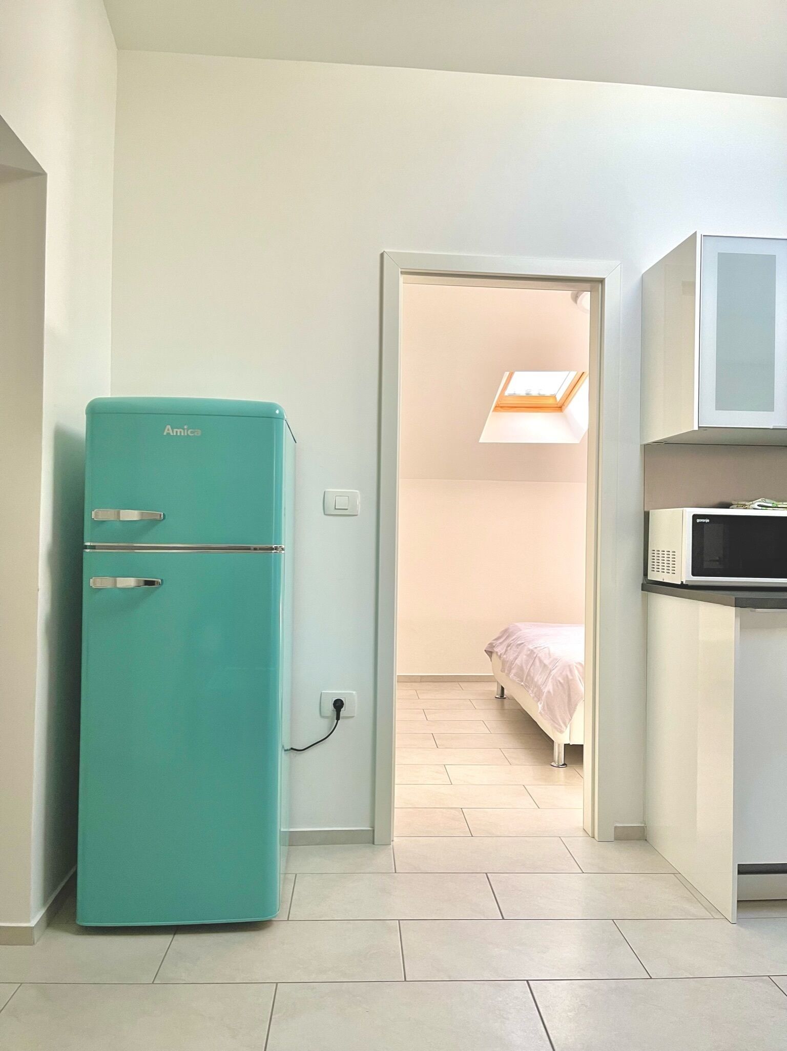 Een kleine keuken met een blauwgroene koelkast en een magnetron, met een bed in de aangrenzende kamer.