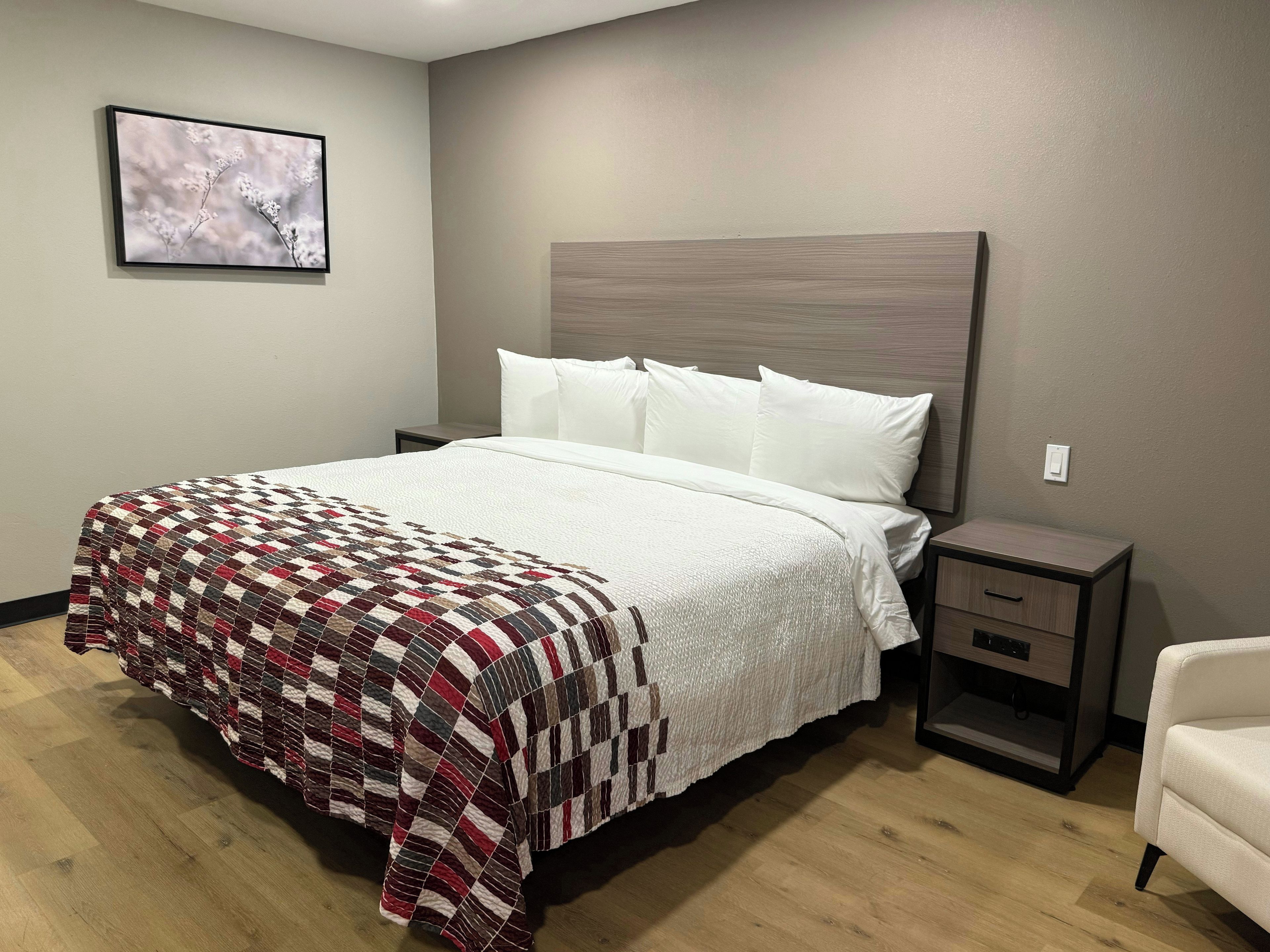 A neatly made bed with a quilt, a bedside table with drawers, a chair, and a framed picture on the wall.