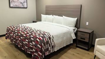A neatly made bed with a quilt, a bedside table with drawers, a chair, and a framed picture on the wall.