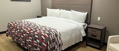 A neatly made bed with a quilt, a bedside table with drawers, a chair, and a framed picture on the wall.