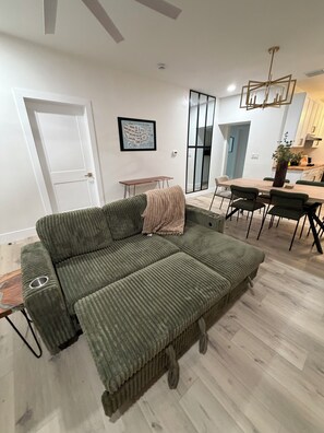 Een woonkamer met een groene bank, een eettafel met stoelen, een keuken en een witte deur.