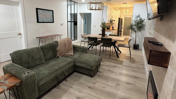 Un salon moderne avec un canapé d’angle, un coin repas avec des chaises, une cuisine en arrière-plan et un ventilateur de plafond.
