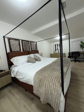 Een slaapkamer met een hemelbed, een bureau met stoel, een bloempot en kunst aan de muur.