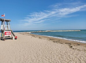 A lifeguard tower with a red flag, a clear blue sky, and a sandy beach with a rocky breakwater in the distance. - Ca Festa Residence: Two-room apartment Ca Festa 51 for 6 people (Chioggia)