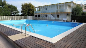 A rectangular swimming pool with a wooden deck surrounding it, a metal ladder, and a modern building with large windows in the background. - Ca Festa Residence: Two-room apartment Ca Festa 51 for 6 people (Chioggia)