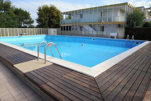 A rectangular swimming pool with a wooden deck surrounding it, a metal ladder, and a modern building with large windows in the background. - Ca Festa Residence: Two-room apartment Ca Festa 51 for 6 people (Chioggia)