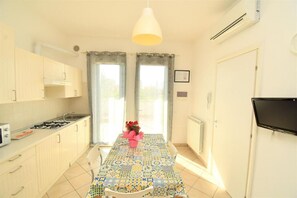 A kitchen with a tiled floor, a gas stove, cabinets, a dining table with a patterned tablecloth, and a TV mounted on the wall. - Residenz Arcobaleno, Stella Marina 6 Two-Room Apartment for 4 People (Chioggia)