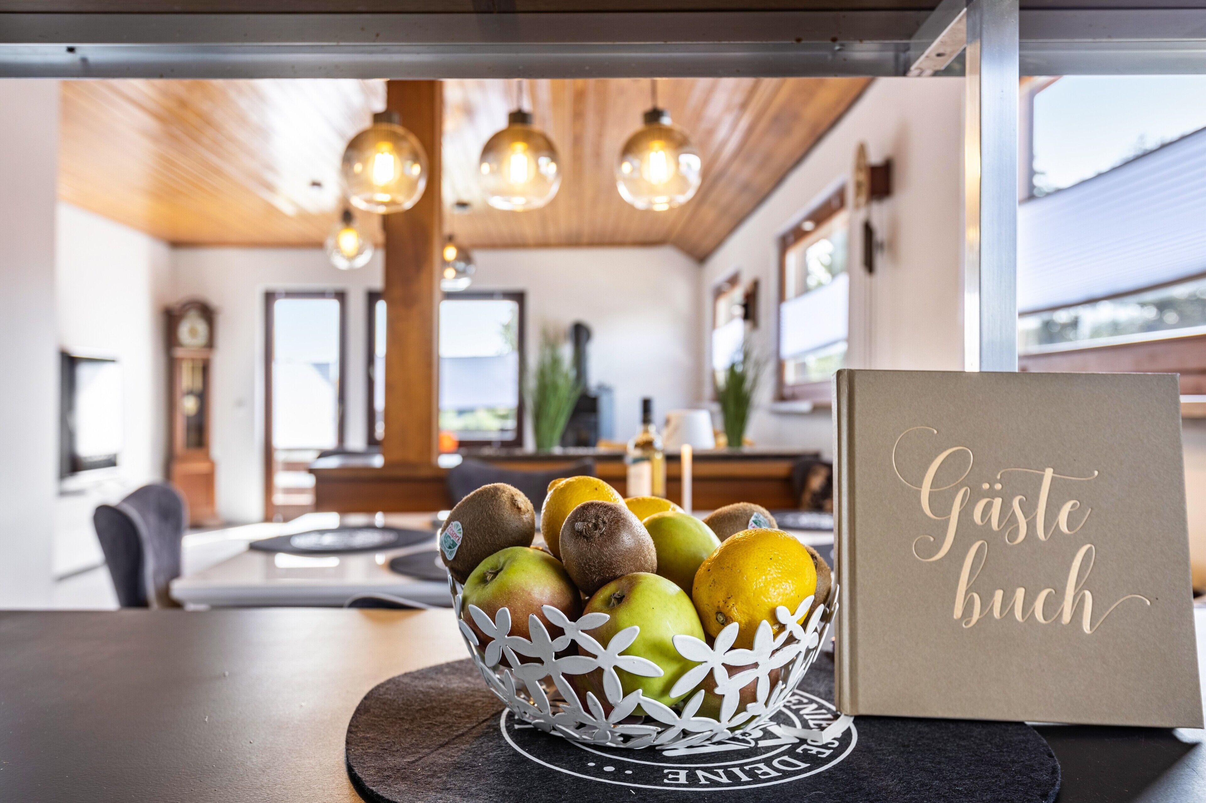A fruit bowl with apples and kiwis on a coaster, a card with 'Gäste buch' text, and a modern interior with wooden ceiling and pendant lights.