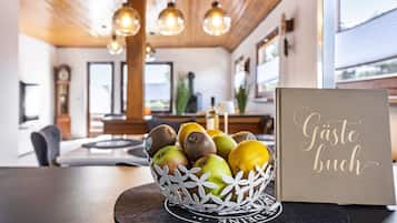 Uma tigela de frutas com maçãs e kiwis sobre um apoio, um cartão com o texto 'Gäste buch', e um interior moderno com teto de madeira e luminárias pendentes.