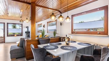A modern dining area with a wooden ceiling, pendant lighting, and large windows offering views of the outdoors.