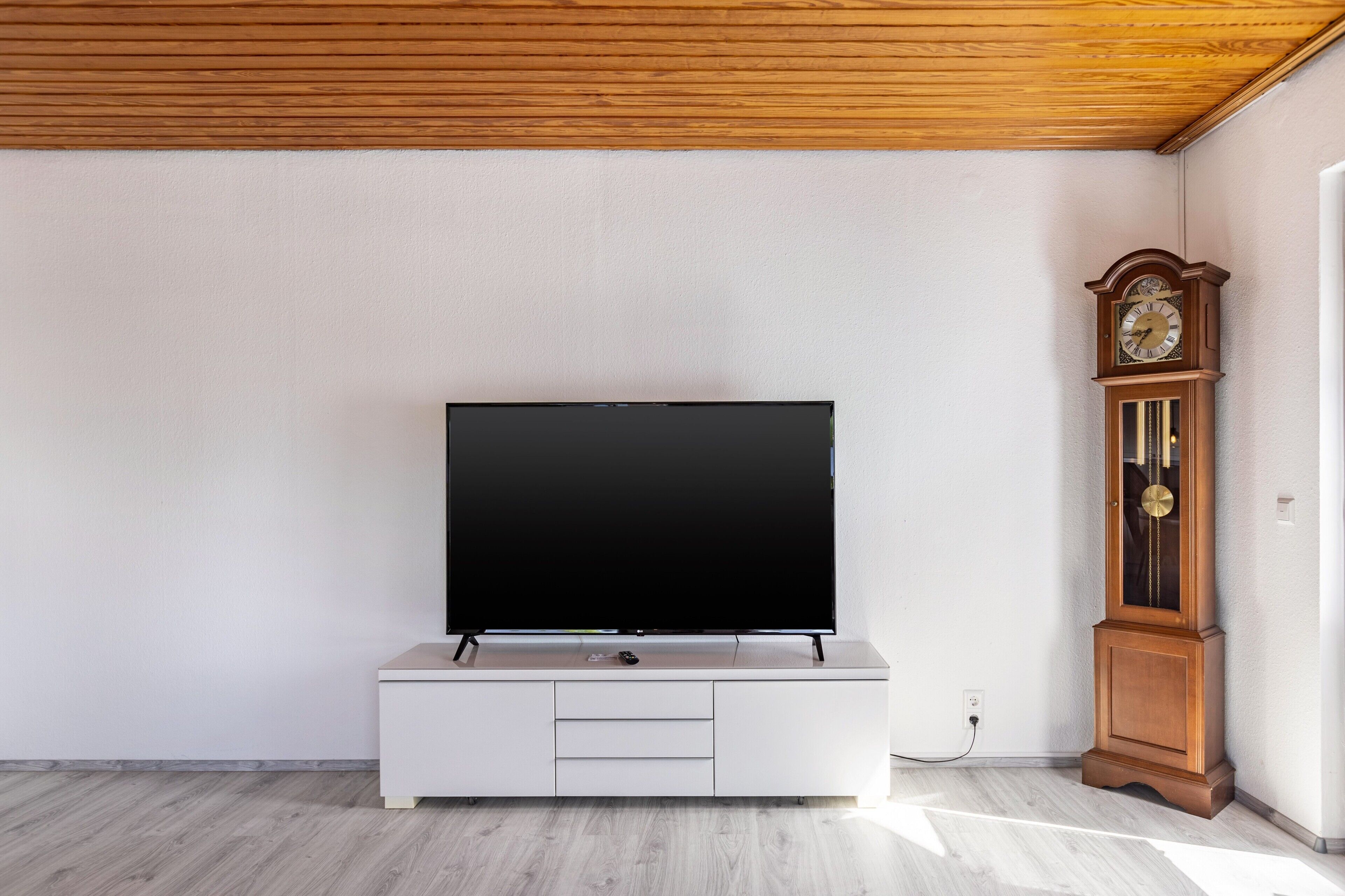 Un salon moderne avec un téléviseur à écran plat posé sur un meuble blanc, un plafond en bois et une horloge fixée au mur.