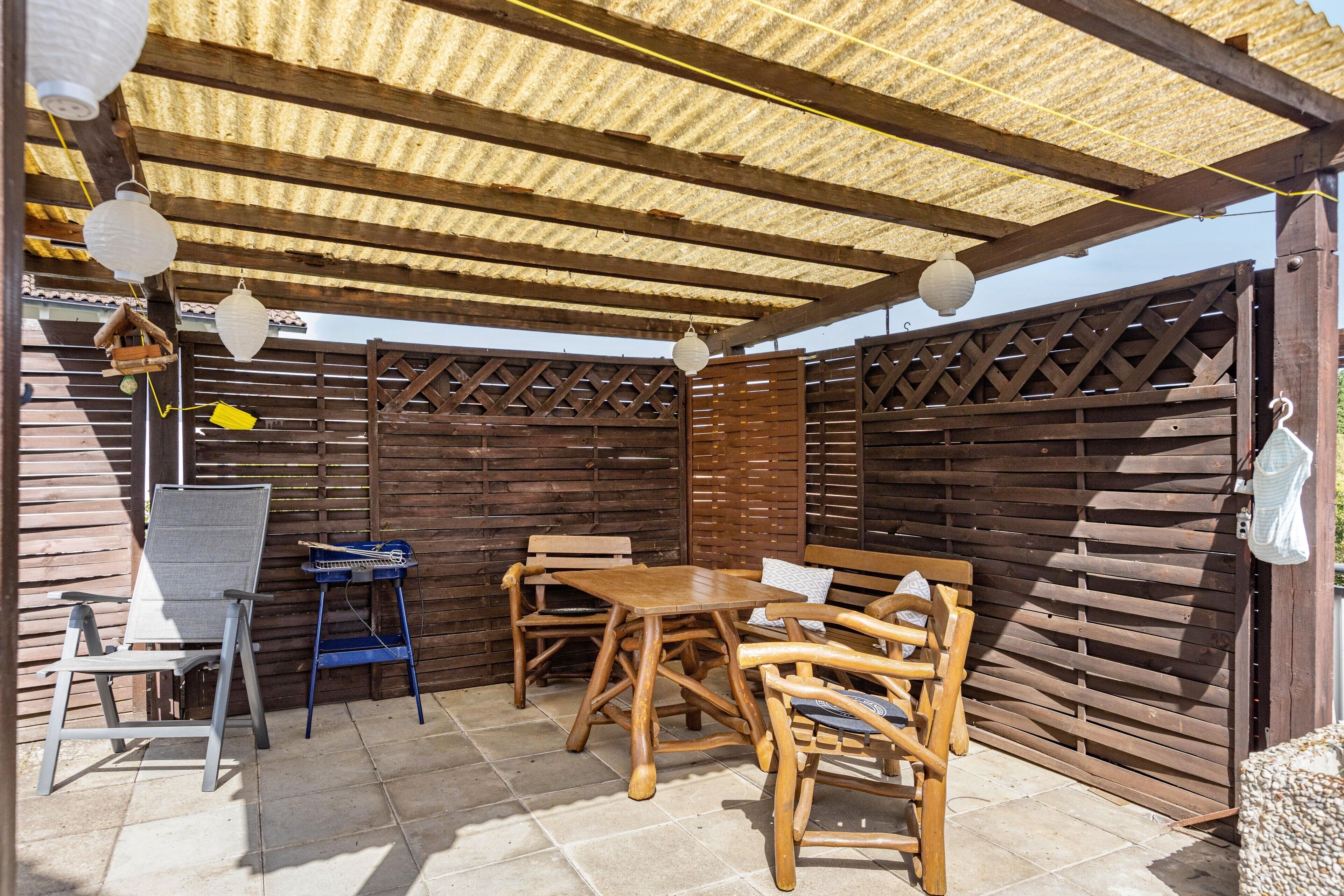 Eine Terrasse im Freien mit einem Holztisch und Stühlen, einem blauen Klappstuhl und einem weißen Laternenhängeleuchter.