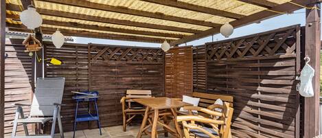 Eine Terrasse im Freien mit einem Holztisch und Stühlen, einem blauen Klappstuhl und einem weißen Laternenhängeleuchter.