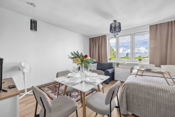 Moderni hotellihuone, jossa on ruokailutila, sänky, työpöytä ja suuri ikkuna, josta avautuu näkymä kaupunkiin.