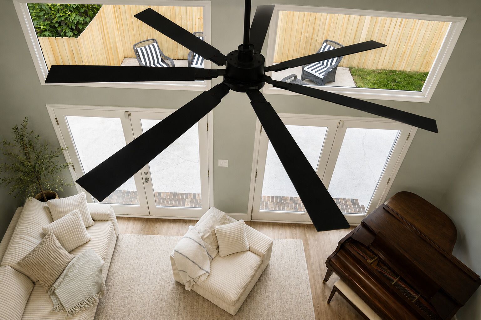 Una habitación con ventilador de techo, un piano, un sofá y vistas a un patio con sillas y valla de madera.