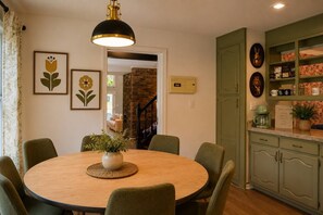 Ein Essbereich mit einem runden Tisch, Stühlen, einer Hängelampe und einer Küchenzeile mit Schränken und einer Kaffeemaschine.