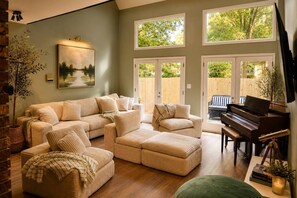 A living room with a sectional sofa, a piano, and a large window with a view of trees. - Sleeps 19 | 2 acres | Pets | MidCity | Space Camp (Huntsville)