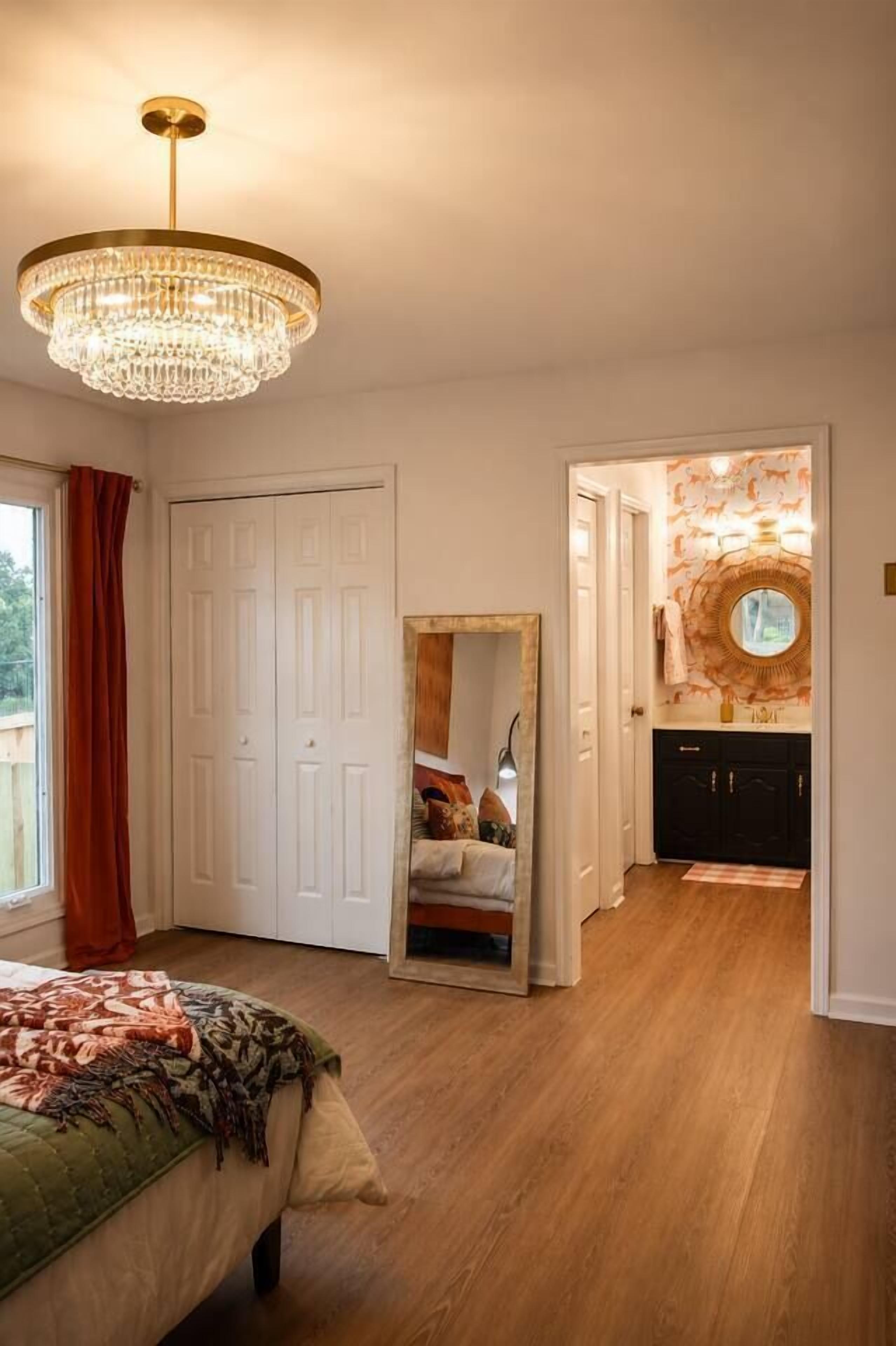 A bedroom with a bed, a chandelier, a mirror, and a bathroom with a round mirror and sink.