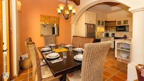 Un coin repas avec une table en bois dressée pour quatre personnes, entourée de chaises en osier, d’un lustre et d’une cuisine en arrière-plan.