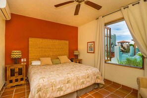 Ein Schlafzimmer mit einem großen Bett, einem hölzernen Kopfteil, zwei Nachttischlampen, einem Fenster mit Vorhängen und Blick auf einen Pool und Gebäude im Freien.