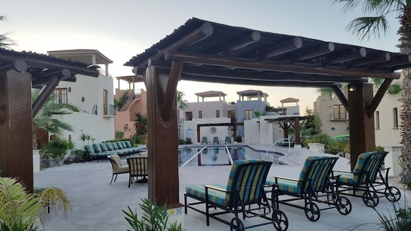 Zona de piscina con sillones de descanso, una pérgola y vistas a los edificios del complejo.