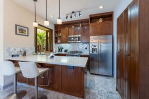 Una cocina con armarios de madera, un refrigerador de acero inoxidable, un microondas y una cafetera. Hay una barra con dos sillas blancas.