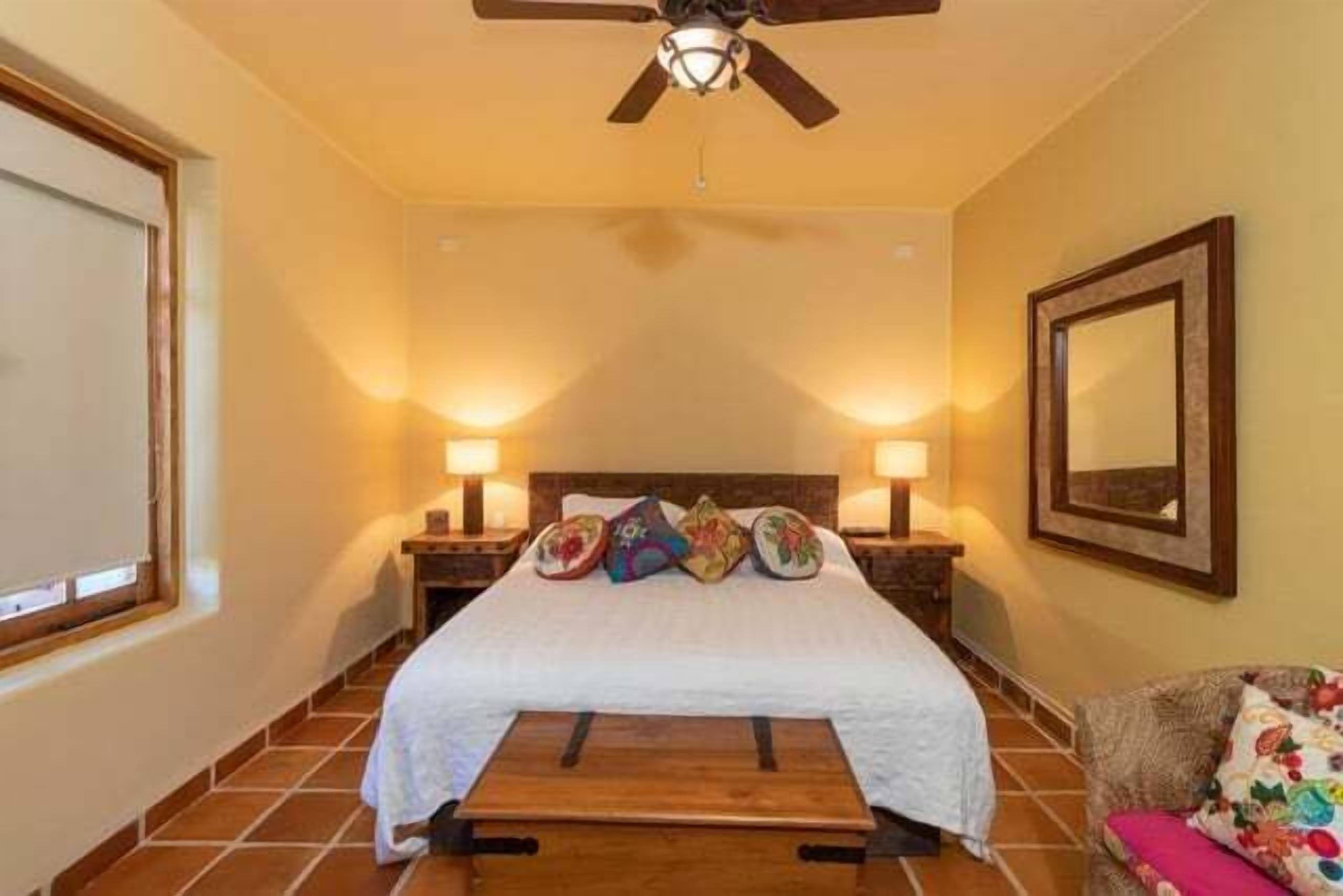 A bedroom with a bed, two bedside lamps, a ceiling fan, a wooden bench, a chair with a cushion, and a framed mirror on the wall.