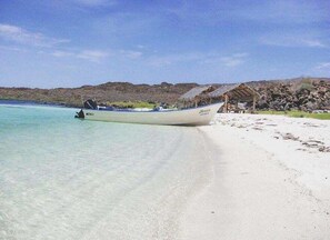 A boat on a sandy beach with clear water, a thatched-roof structure, and rocky hills in the background. - Beautiful Custom Home. Amazing Ocean View (  Loreto)