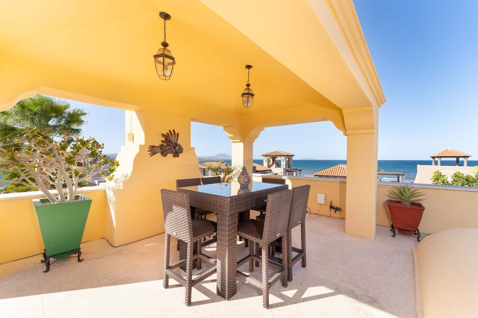A covered outdoor patio with a table and chairs, potted plants, and a view of the sea.