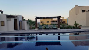 Une piscine entourée de bâtiments, avec des chaises longues et une pergola.