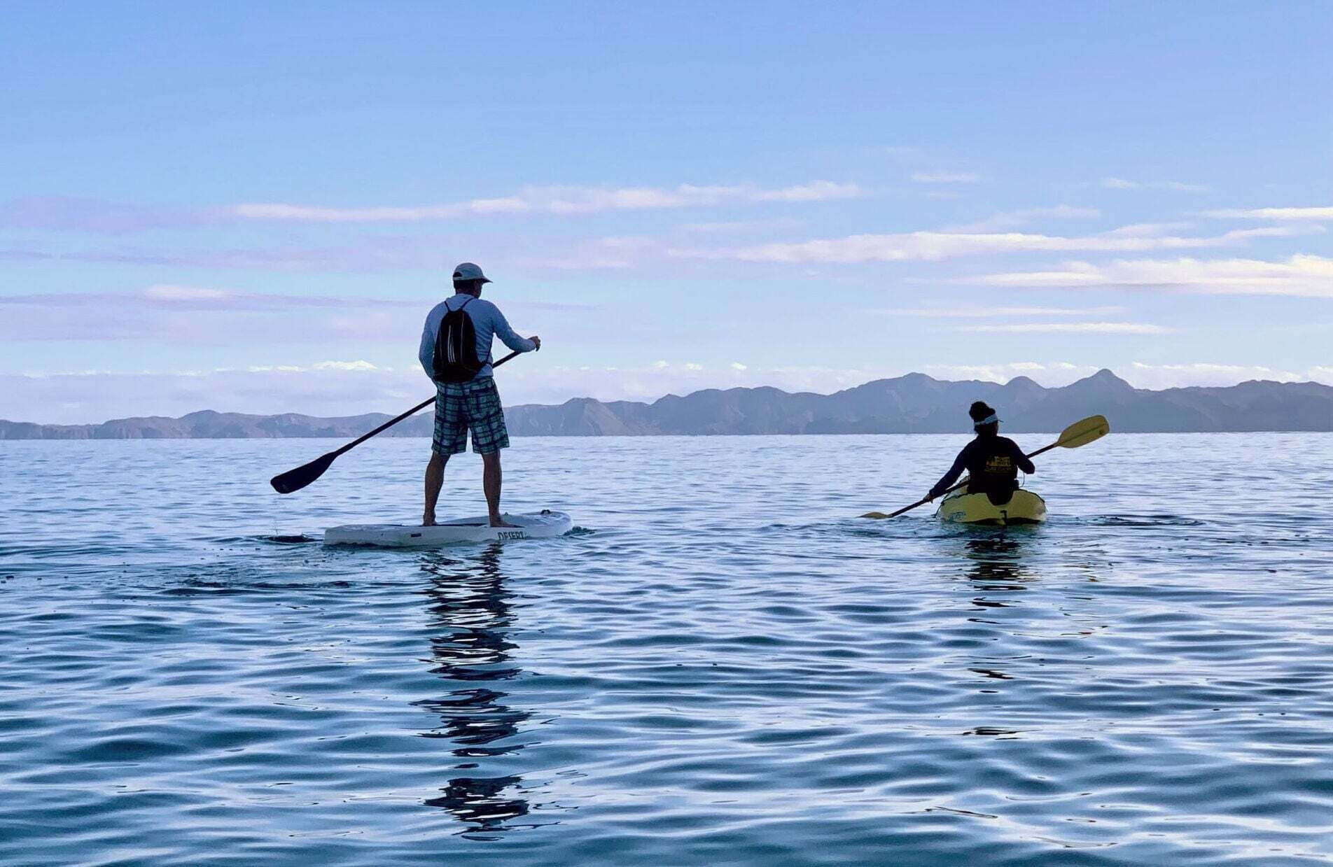 Duas pessoas em águas calmas, uma em pé num stand-up paddle e outra num caiaque, com montanhas ao fundo.