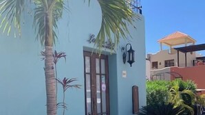 A building with a light blue exterior, a wooden door, and a small porch area with a palm tree and potted plants. - Discover This Charming 2-bedroom Home Located in Founders Area! (  Loreto)
