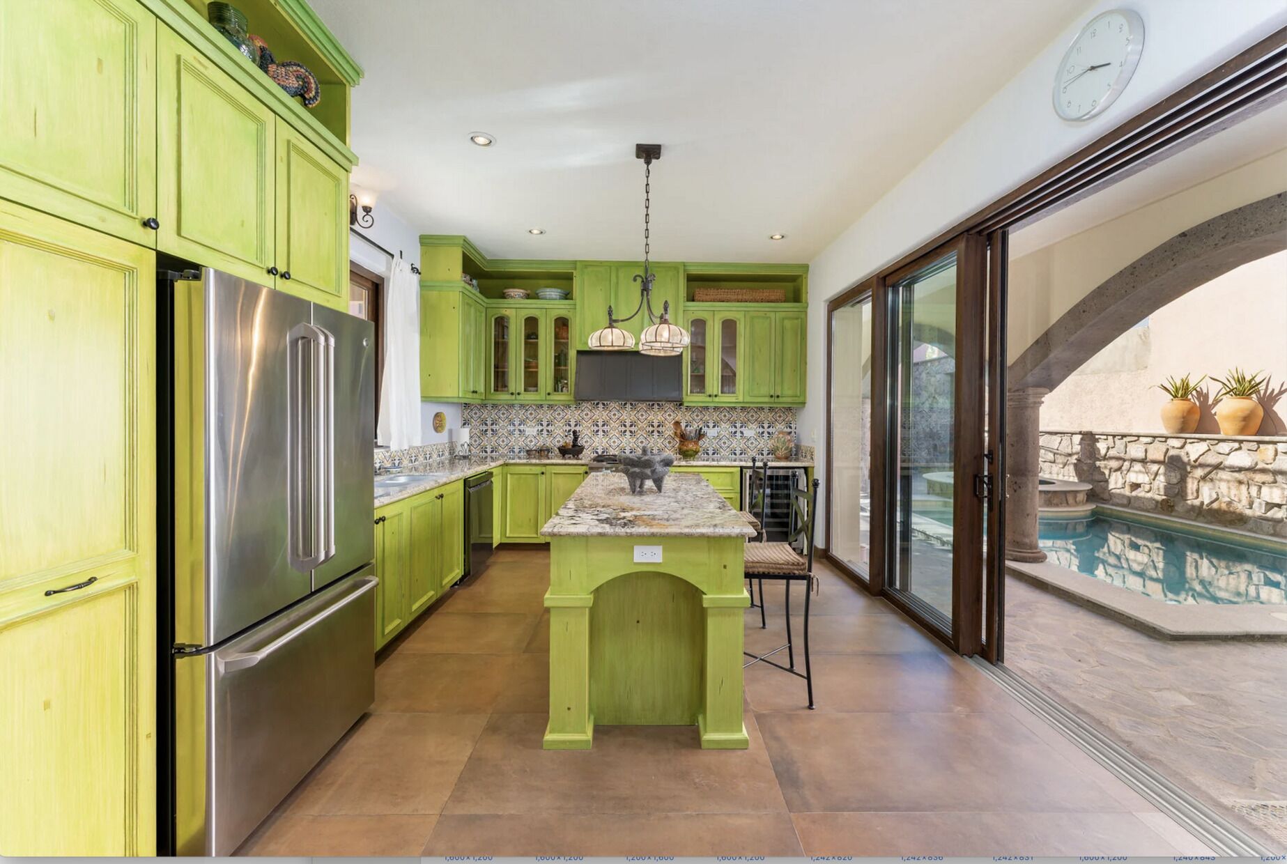 Cocina con armarios verdes, un refrigerador de acero inoxidable, salpicadero de azulejos y vistas a una piscina al aire libre.